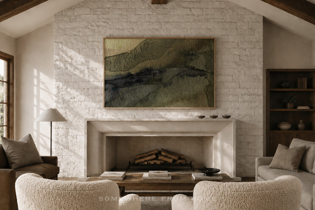 A framed abstract landscape painting with green, beige, and dark blue-gray brushstrokes hangs on a white brick fireplace wall in a cozy living room with textured cream armchairs, a coffee table, and a fireplace.