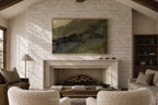 A framed abstract landscape painting with green, beige, and dark blue-gray brushstrokes hangs on a white brick fireplace wall in a cozy living room with textured cream armchairs, a coffee table, and a fireplace.