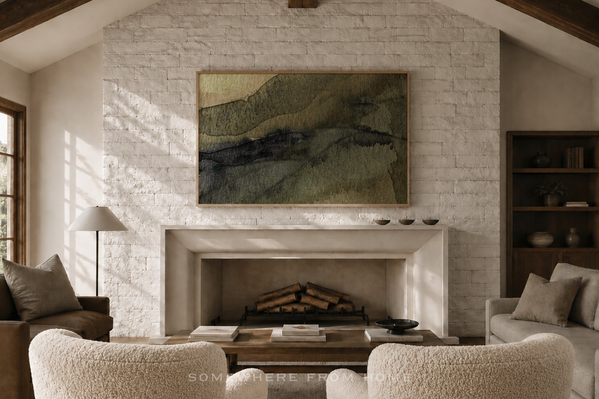 A framed abstract landscape painting with green, beige, and dark blue-gray brushstrokes hangs on a white brick fireplace wall in a cozy living room with textured cream armchairs, a coffee table, and a fireplace.
