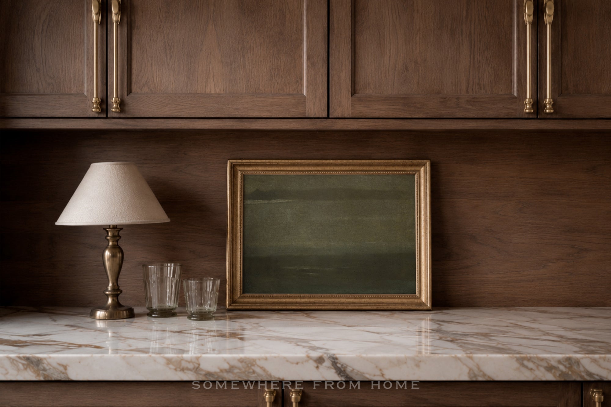 A framed landscape art print with a gold frame and muted green landscape, styled on a marble countertop next to a table lamp and two glasses in a warm, dark wood kitchen setting.