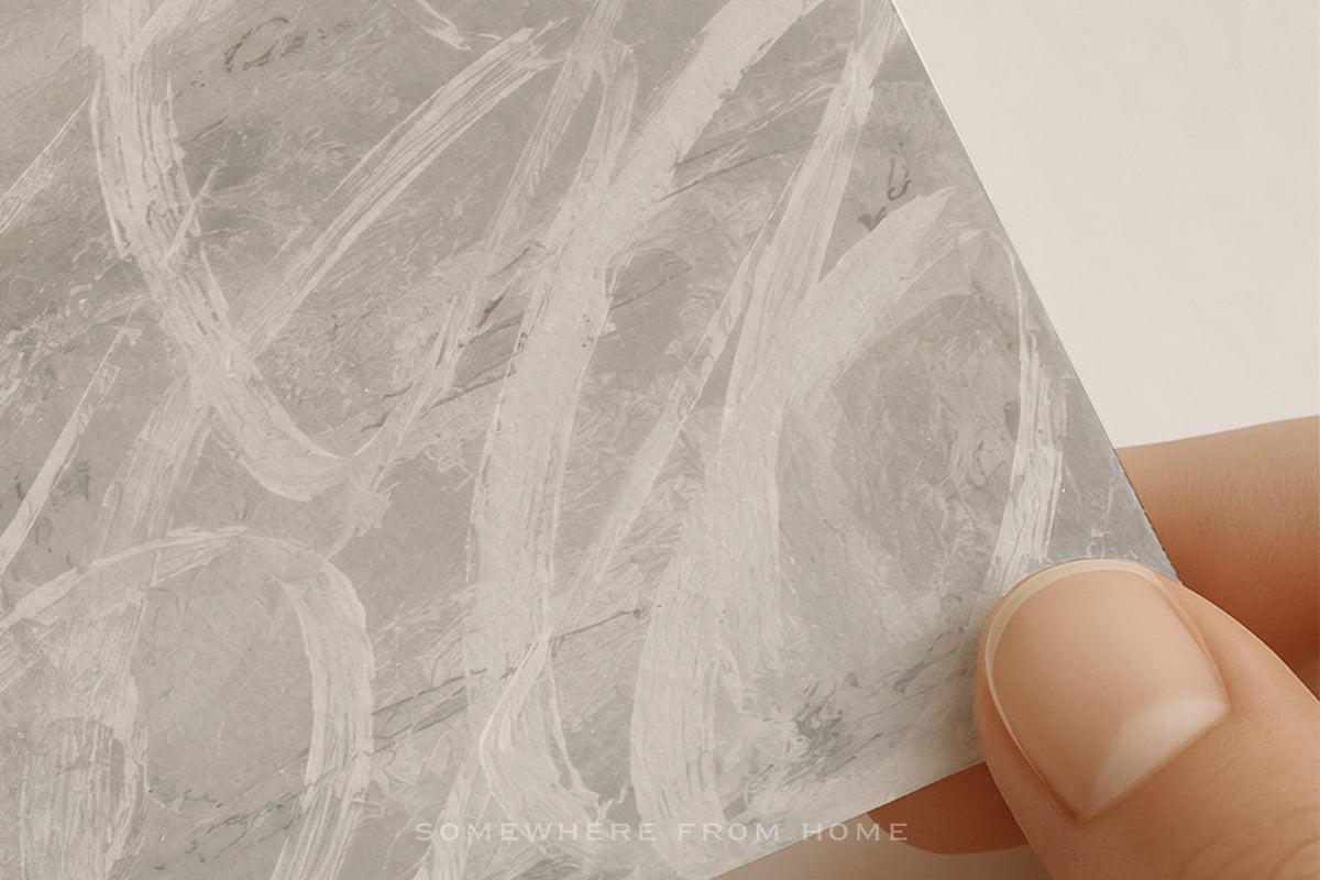 A close-up of a hand holding a square sample of gray marble-patterned wallpaper or adhesive contact paper with white hand-painted style brushstrokes. The brand name 'SOMETHING FROM HOME' is visible at the bottom.