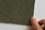 A close-up of a hand holding the edge of a textured dark olive green textured matte art print with a torn raw edge, set against a plain white background. A faint watermark at the bottom reads 'SOMEWHERE FROM HOME'.