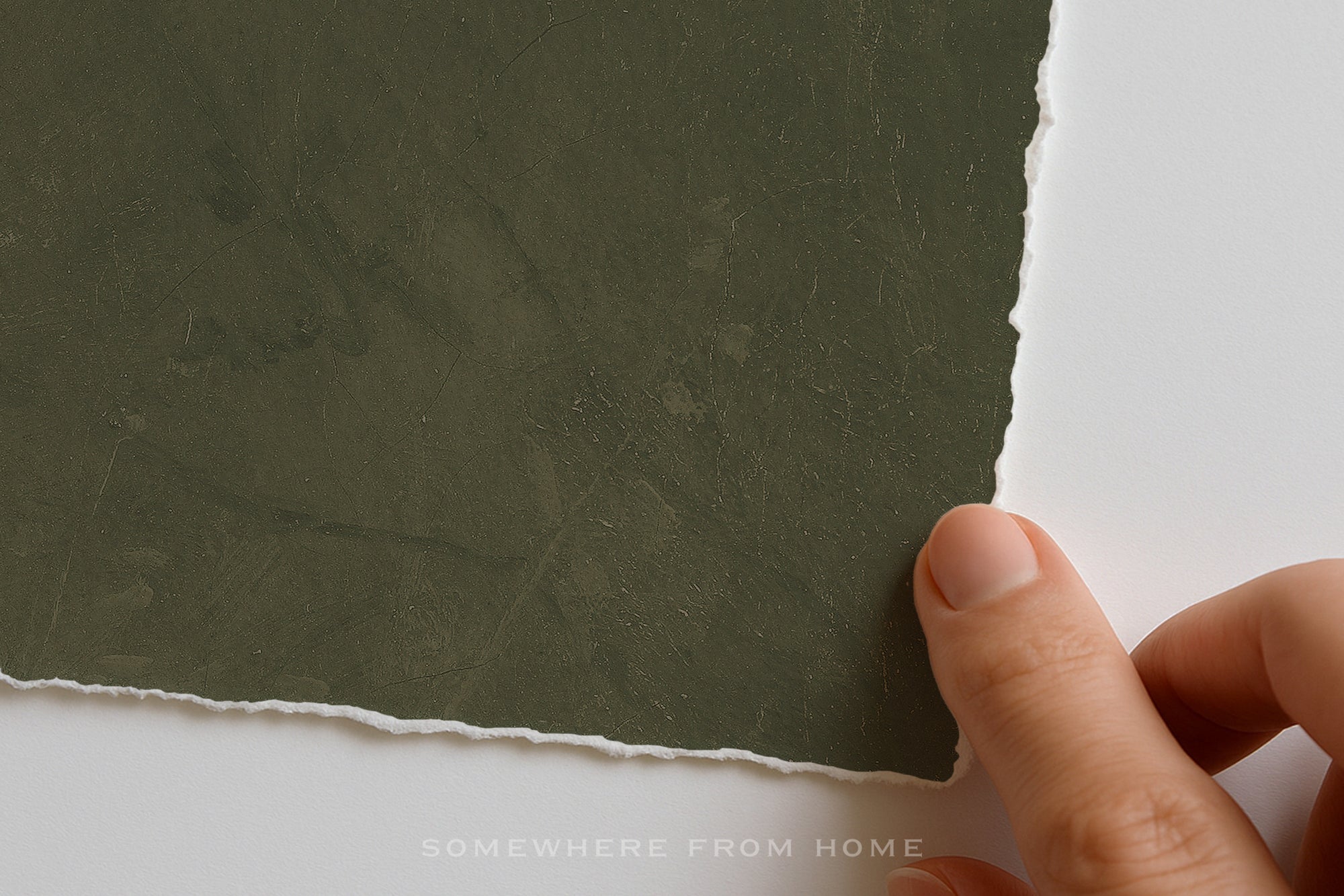 A close-up of a hand holding the edge of a textured dark olive green textured matte art print with a torn raw edge, set against a plain white background. A faint watermark at the bottom reads 'SOMEWHERE FROM HOME'.