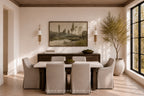 A styled dining room interior featuring a rectangular dining table with six upholstered chairs, a dark wood sideboard, a framed landscape painting on the wall, wall sconces, a large potted tree, and a tall window with black frames.