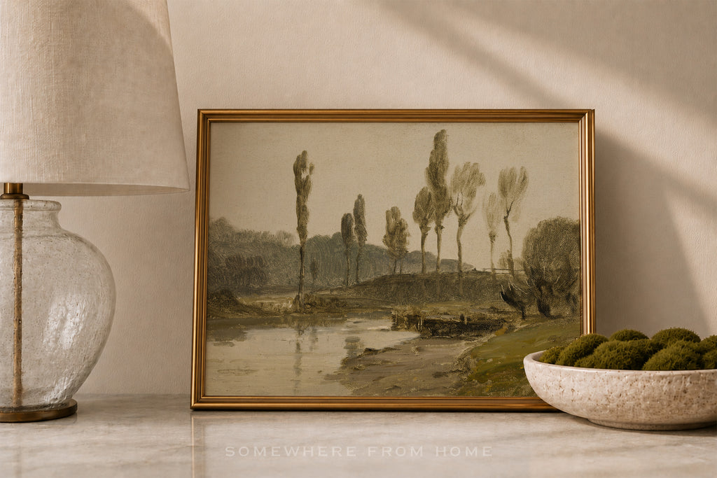 A framed landscape art print depicting tall trees by a river, styled on a marble surface next to a glass lamp and a bowl of green moss.