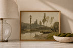 A framed landscape art print depicting tall trees by a river, styled on a marble surface next to a glass lamp and a bowl of green moss.
