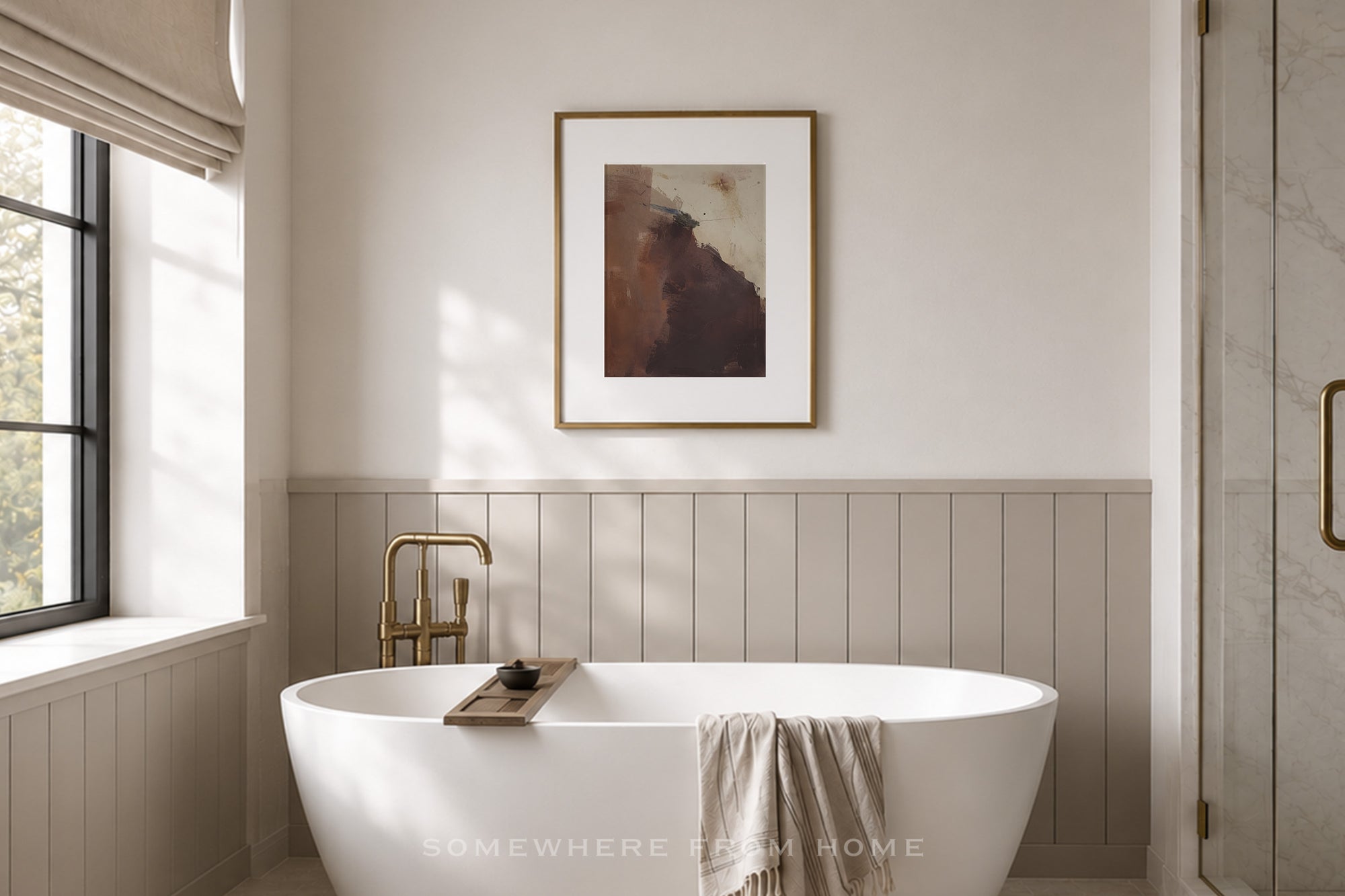 A modern bathroom interior with a white freestanding bathtub, gold faucet, paneled lower wall, and a framed abstract artwork centered above the tub. A window with a beige blind is on the left, and a glass shower door is visible on the right.