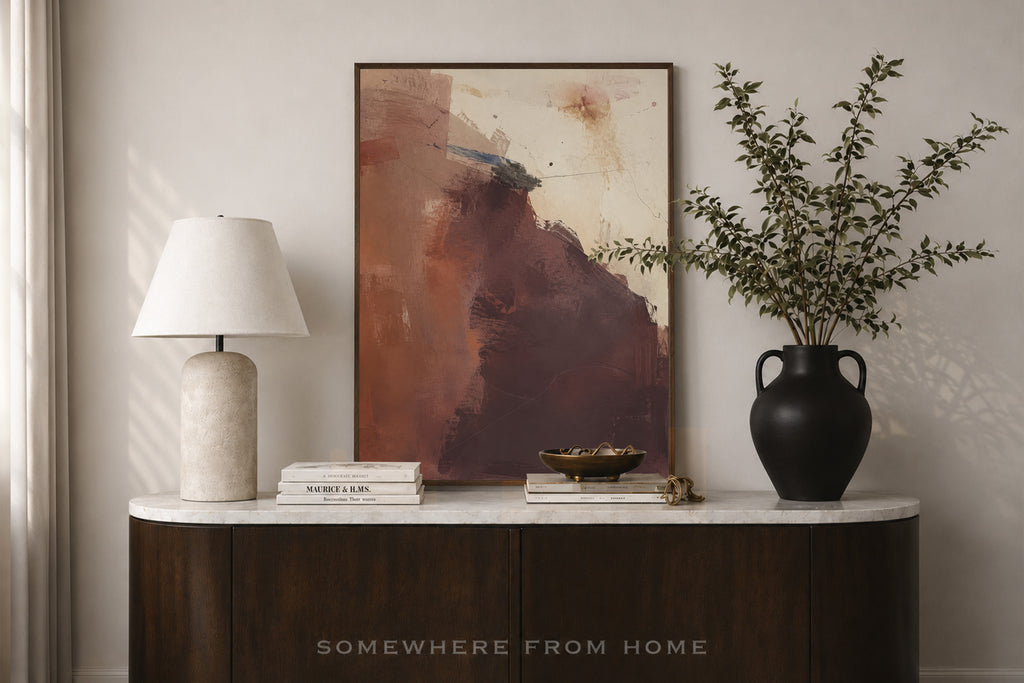 An abstract framed painting with earthy brown, terracotta, and cream tones displayed on a white marble-top console table in a styled interior. A white table lamp, stacked books, a small bowl, and a black vase with leafy branches sit beside it.
