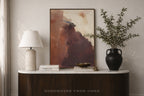 An abstract framed painting with earthy brown, terracotta, and cream tones displayed on a white marble-top console table in a styled interior. A white table lamp, stacked books, a small bowl, and a black vase with leafy branches sit beside it.