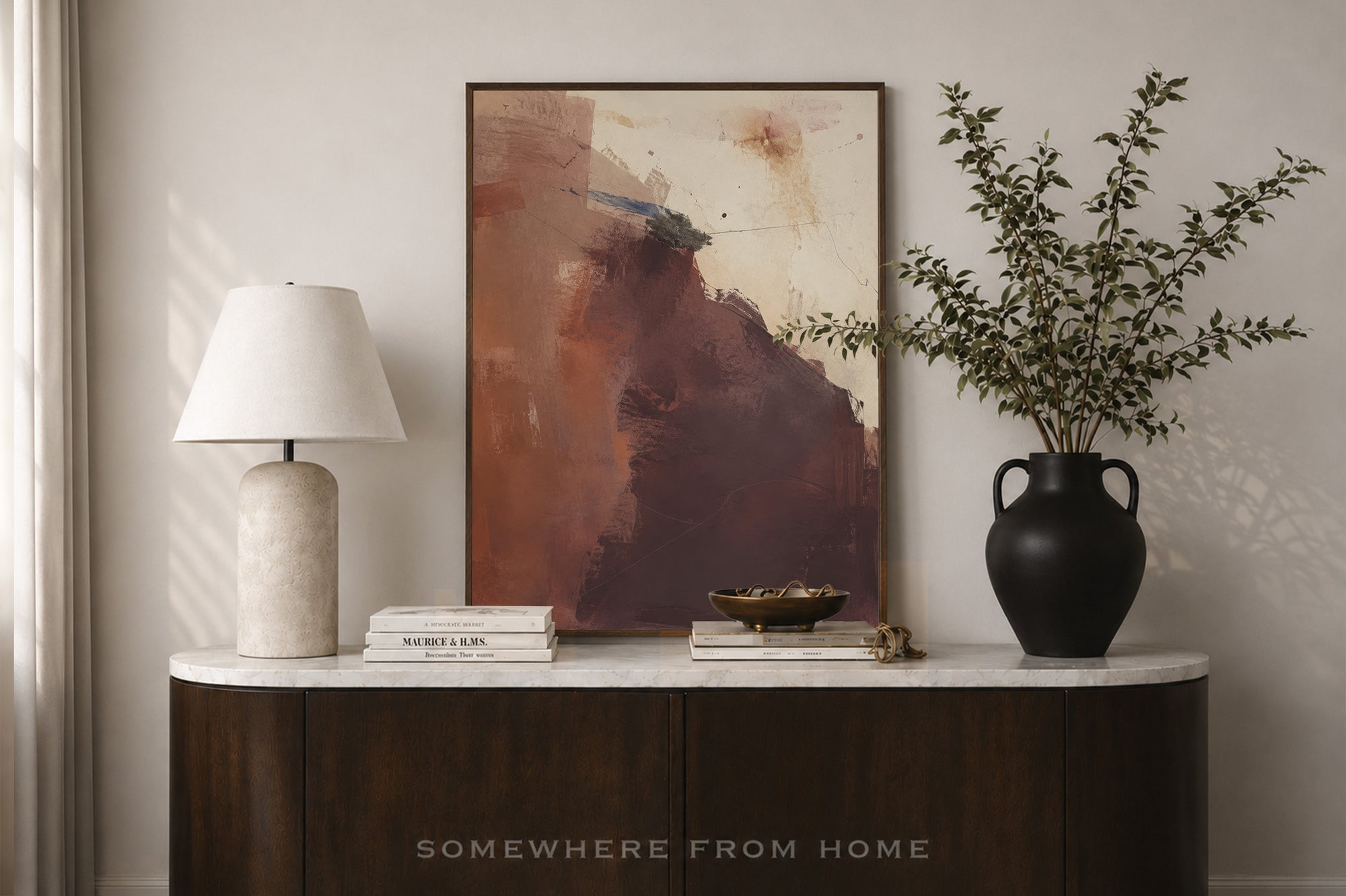 An abstract framed painting with earthy brown, terracotta, and cream tones displayed on a white marble-top console table in a styled interior. A white table lamp, stacked books, a small bowl, and a black vase with leafy branches sit beside it.