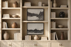 A built-in cream-colored bookshelf with open shelves and lower cabinets, styled with books, ceramic vases, bowls, and two black-and-white landscape framed prints. A watermark reading 'SOMEWHERE FROM HOME' is visible at the bottom.