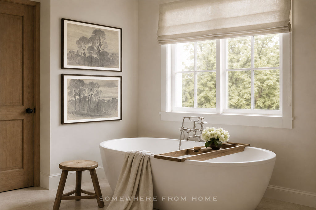 A modern freestanding white bathtub in a bright, neutral-toned bathroom with a window, beige roman shade, wooden stool, and two framed landscape prints on the wall. A wooden tray with white flowers rests on the tub edge.