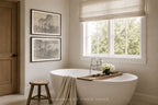 A modern freestanding white bathtub in a bright, neutral-toned bathroom with a window, beige roman shade, wooden stool, and two framed landscape prints on the wall. A wooden tray with white flowers rests on the tub edge.