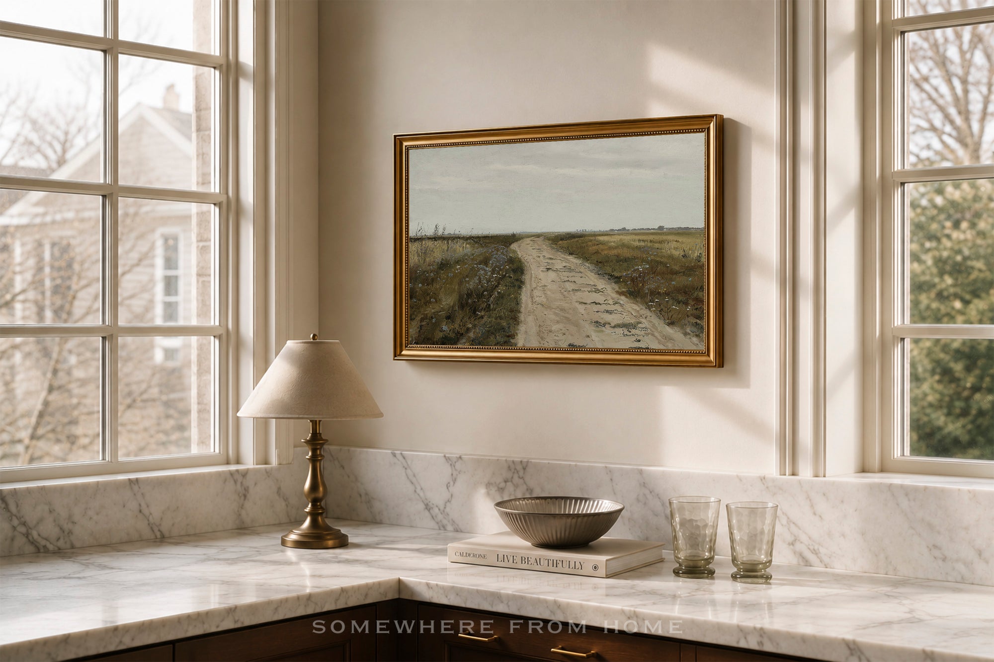 A framed landscape painting of a dirt road through a field hangs on a beige wall above a marble kitchen counter with a table lamp, bowl, and glasses.