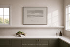 A framed black and white landscape art print hangs on a light beige wall above a white shelf in a modern kitchen setting with dark green cabinets and white countertops.