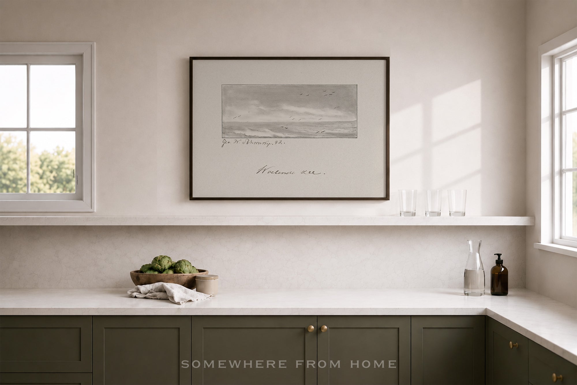 A framed black and white landscape art print hangs on a light beige wall above a white shelf in a modern kitchen setting with dark green cabinets and white countertops.