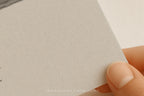 A close-up of a hand holding the corner of a textured light gray paper or card, with a small portion of a printed artwork visible at the top left.