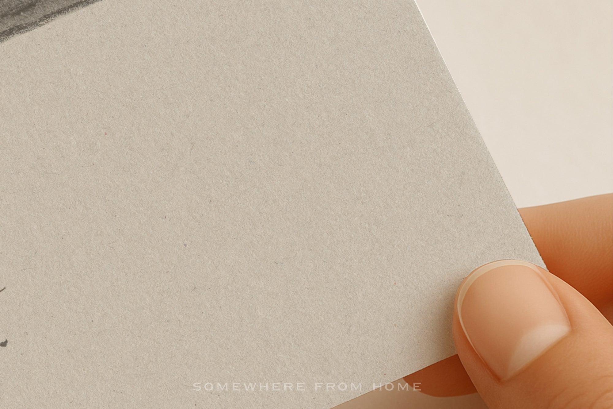 A close-up of a hand holding the corner of a textured light gray paper or card, with a small portion of a printed artwork visible at the top left.