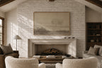 A large framed landscape art print hangs on a white brick fireplace wall in a cozy living room setting with a fireplace, armchairs, and a lamp.