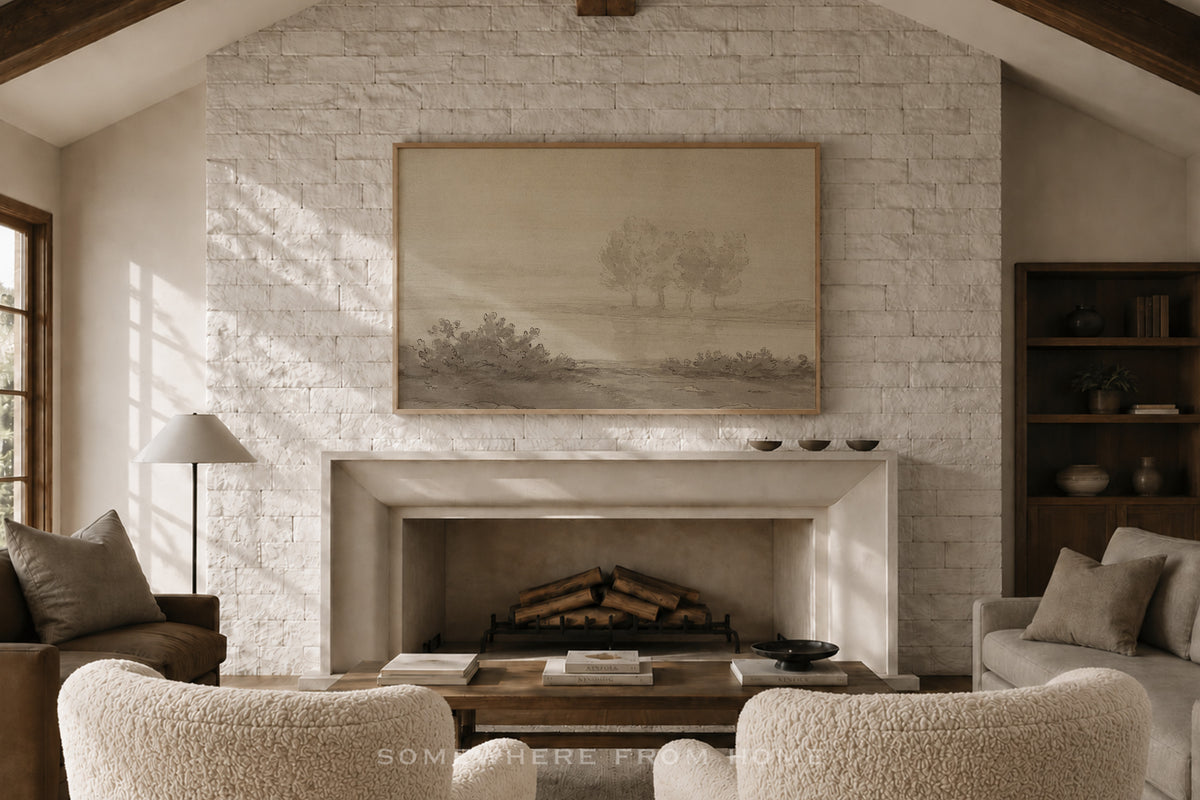 A large framed landscape art print hangs on a white brick fireplace wall in a cozy living room setting with a fireplace, armchairs, and a lamp.