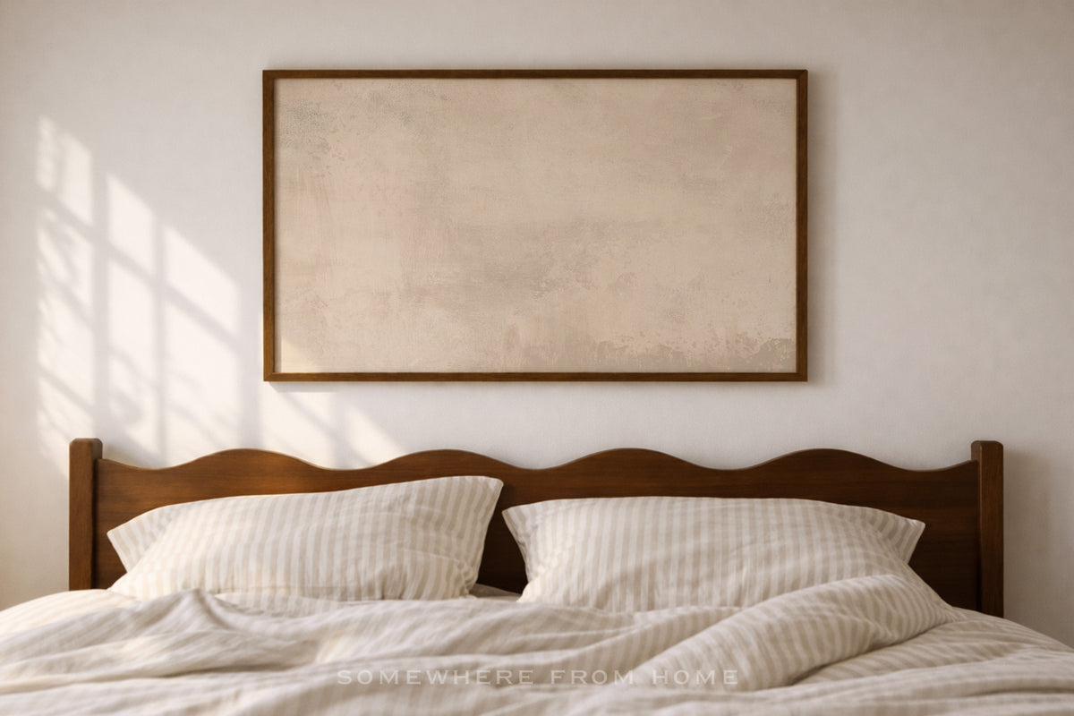 A styled bedroom scene with a wooden bed, cream bedding, two pillows, and a large framed abstract wall art piece hanging above the headboard against a light wall with soft window shadows.