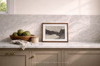 A framed black-and-white landscape photograph in a medium brown wood frame, styled on a white marble kitchen counter with a wooden bowl of artichokes and a beige ceramic container. The background features a matching marble backsplash and beige cabinetry.