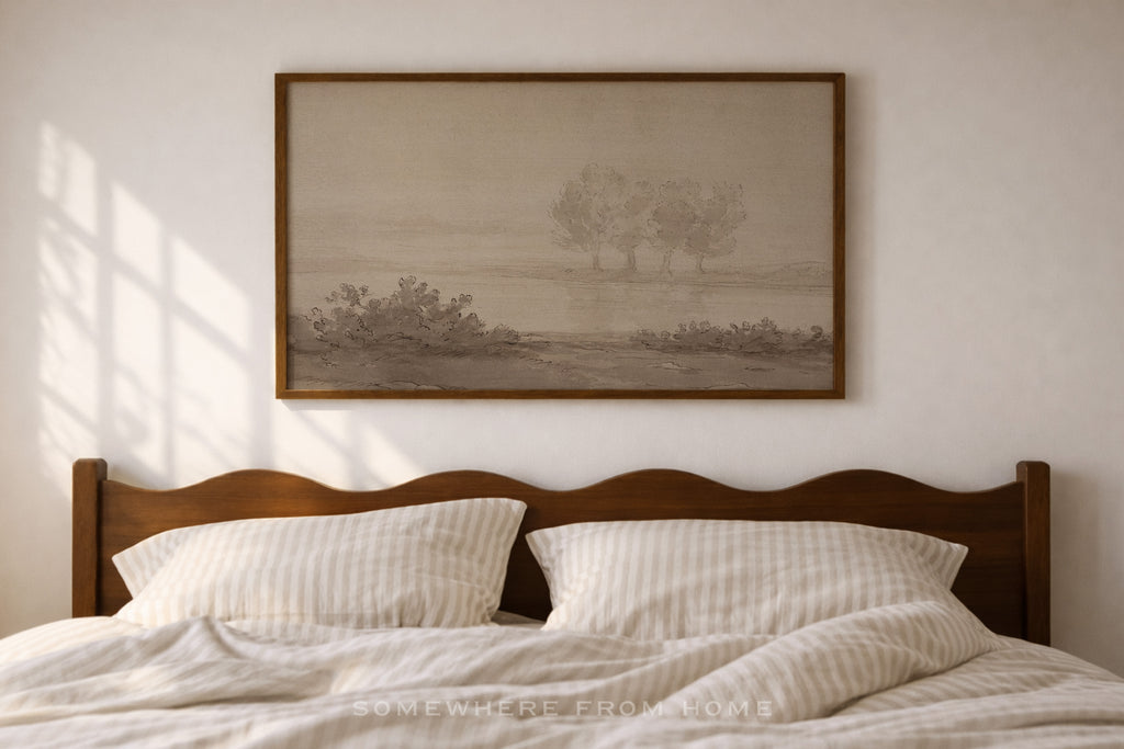 A framed landscape art print featuring two trees in a misty field, hung on a light beige wall above a wooden bed with white striped bedding.
