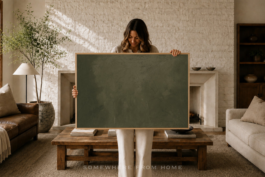 A woman stands in a cozy living room holding a large blank dark green chalkboard with a light wood frame. The room features a white brick fireplace, a wooden coffee table, a leather sofa, and a potted plant in the background.