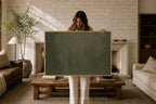 A woman stands in a cozy living room holding a large blank dark green chalkboard with a light wood frame. The room features a white brick fireplace, a wooden coffee table, a leather sofa, and a potted plant in the background.