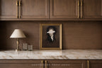 A framed portrait print with a blindfolded figure in dark clothing, displayed on a marble countertop against a dark wood cabinet wall, with a table lamp and two glasses beside it.