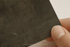 A close-up of a hand holding a dark charcoal textured tile or stone sample against a plain white background.