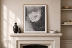 A framed abstract black and white art print with a light wood frame, displayed on a cream-colored stone mantel in a styled room setting with a vase, bowls, and shelves.