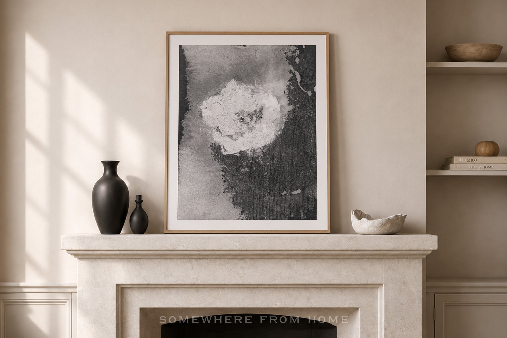 A framed abstract black and white art print with a light wood frame, displayed on a cream-colored stone mantel in a styled room setting with a vase, bowls, and shelves.