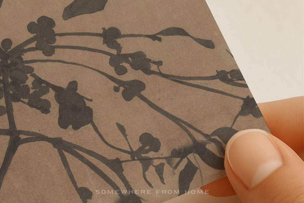 A close-up of a hand holding a square sample of textured kraft paper featuring a dark brown floral branch pattern, with a white background visible in the corner.