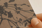 A close-up of a hand holding a square sample of textured kraft paper featuring a dark brown floral branch pattern, with a white background visible in the corner.