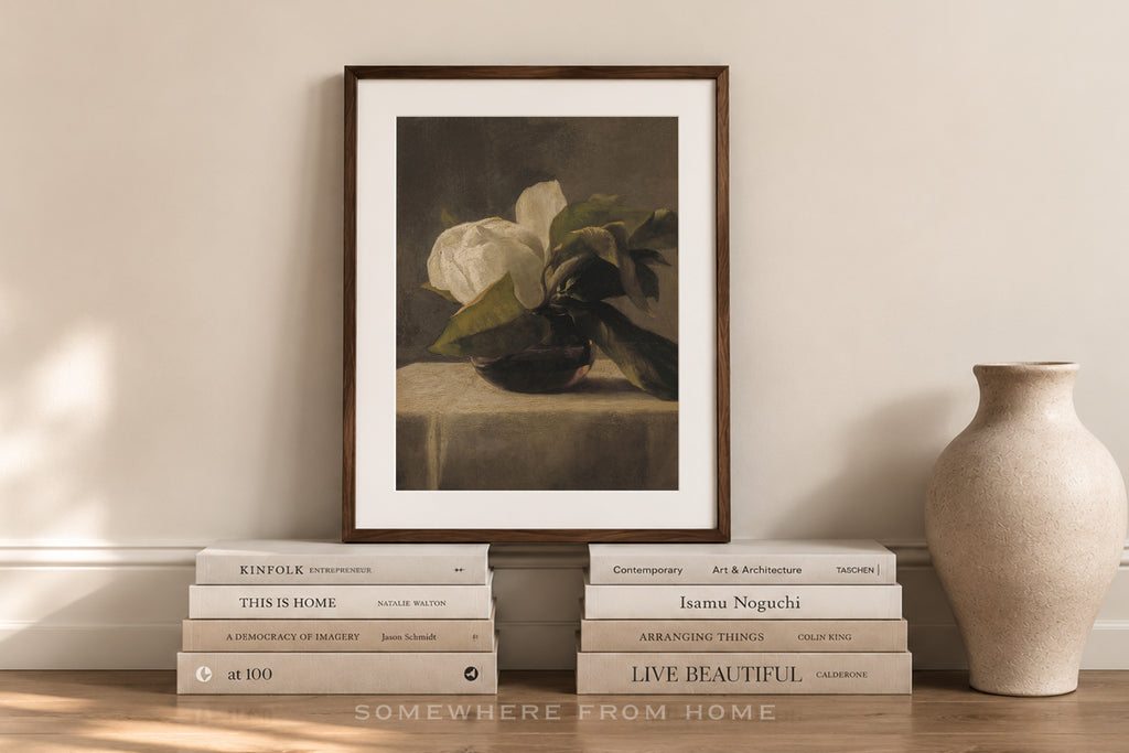 A framed still life art print featuring white flowers in a dark vase, styled on a wooden shelf with stacked books and a ceramic vase against a warm beige wall.