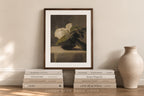 A framed still life art print featuring white flowers in a dark vase, styled on a wooden shelf with stacked books and a ceramic vase against a warm beige wall.