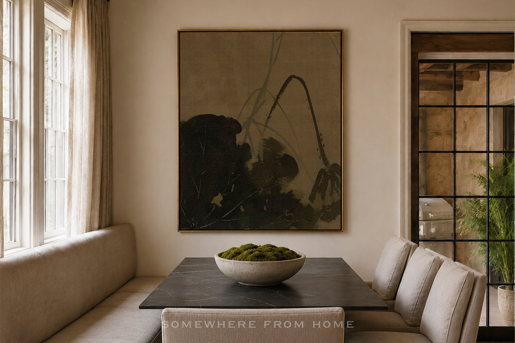 A large framed abstract painting hangs on a beige wall above a dark wooden dining table with upholstered chairs in a softly lit room. The artwork features dark, organic brushstrokes on a muted taupe background.