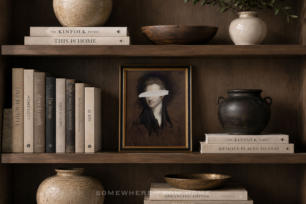 A framed portrait print with a white eye mask over the eyes, displayed on a dark wooden shelf alongside books, ceramic vases, and bowls.