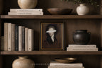 A framed portrait print with a white eye mask over the eyes, displayed on a dark wooden shelf alongside books, ceramic vases, and bowls.