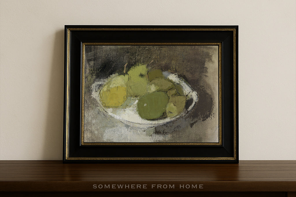 A framed still life painting of green and yellow apples on a white plate, set in a black frame with gold trim, leaning against a beige wall on a dark wooden shelf.