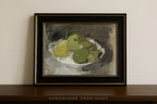 A framed still life painting of green and yellow apples on a white plate, set in a black frame with gold trim, leaning against a beige wall on a dark wooden shelf.