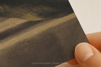 A close-up of a hand holding a printed art card or postcard featuring a textured landscape painting in earthy brown, beige, and dark gray tones. The text 'SOMEWHERE FROM HOME' is printed at the bottom.