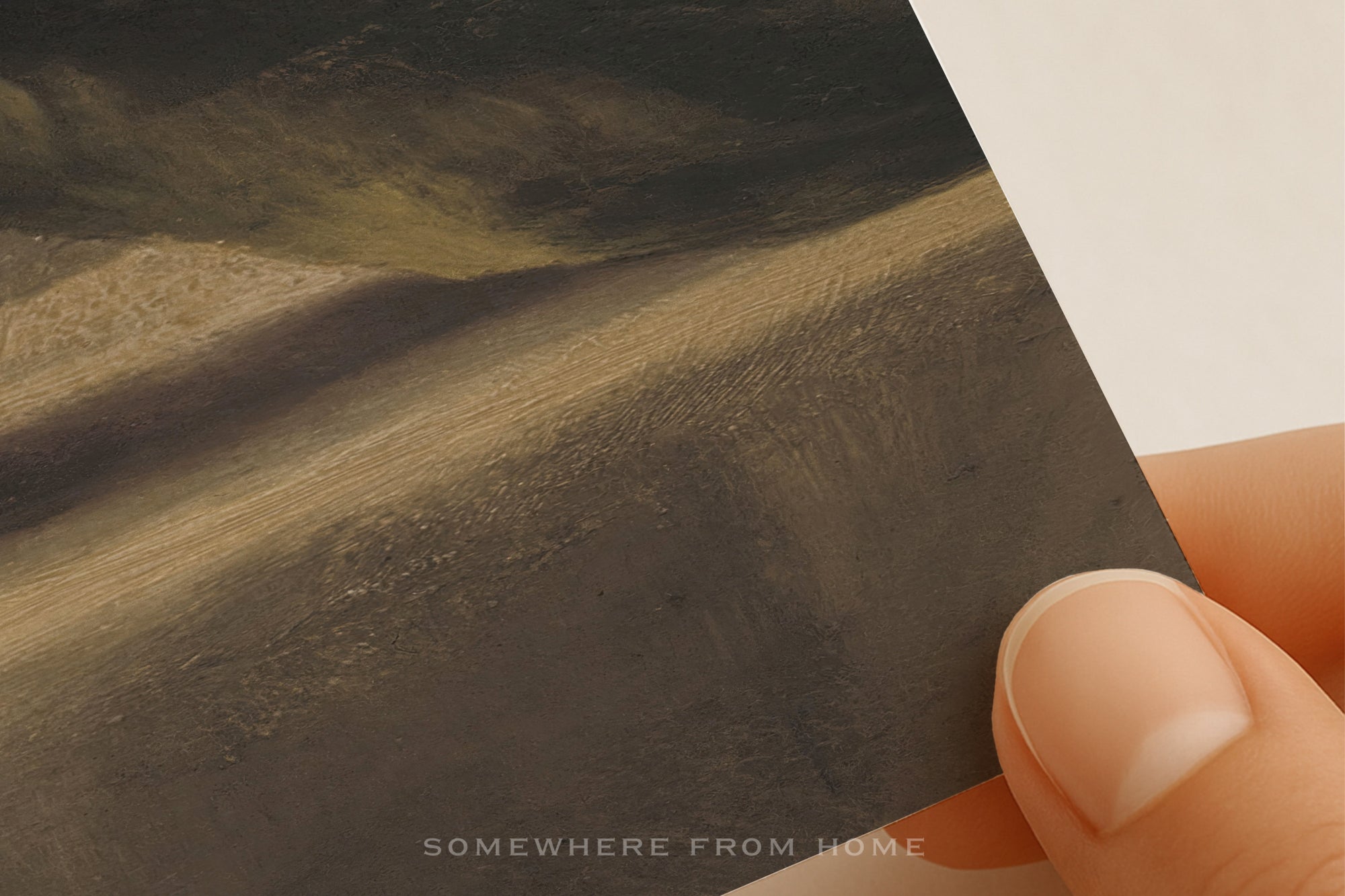 A close-up of a hand holding a printed art card or postcard featuring a textured landscape painting in earthy brown, beige, and dark gray tones. The text 'SOMEWHERE FROM HOME' is printed at the bottom.