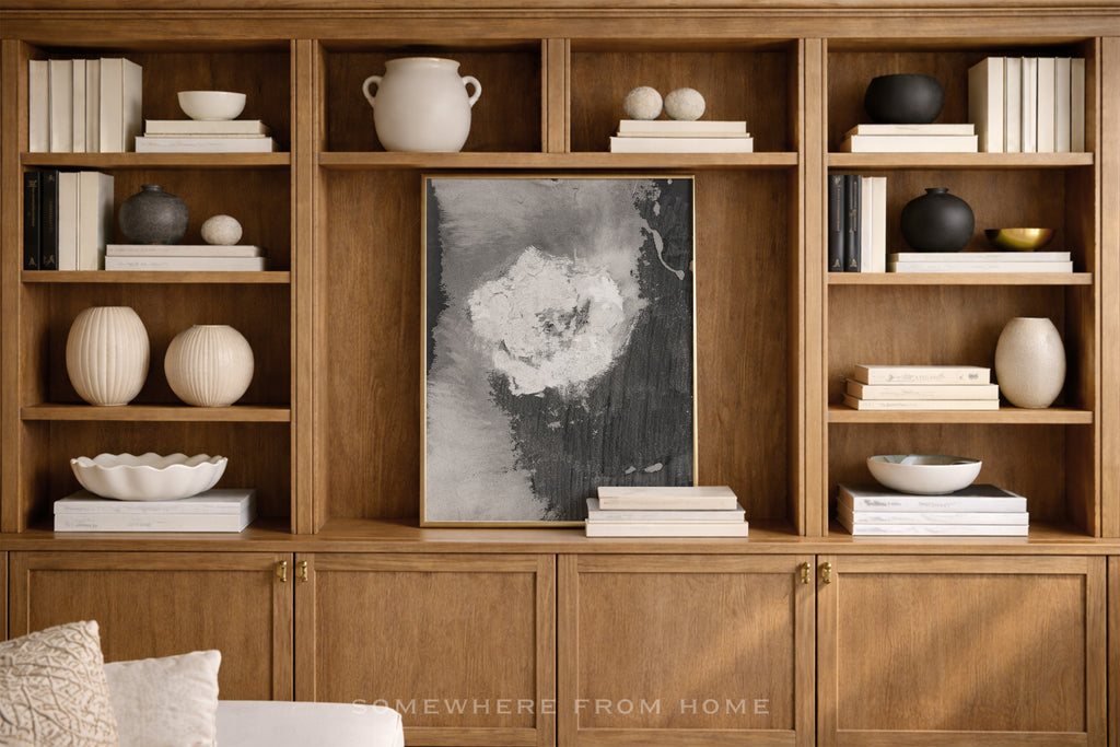 A large wooden built-in bookcase with open shelves and lower cabinet doors, styled with books, ceramic vases, bowls, and a central black-and-white abstract wall art piece. A small 'SOMEWHERE FROM HOME' watermark appears near the bottom.