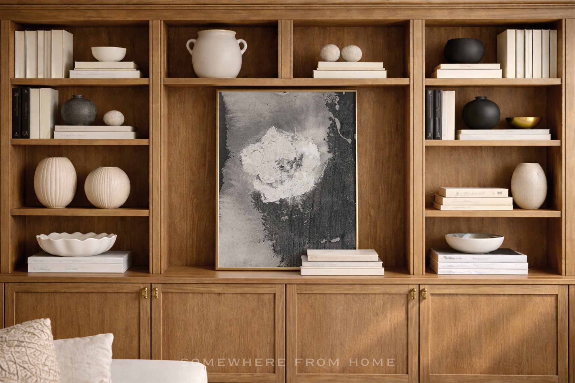 A large wooden built-in bookcase with open shelves and lower cabinet doors, styled with books, ceramic vases, bowls, and a central black-and-white abstract wall art piece. A small 'SOMEWHERE FROM HOME' watermark appears near the bottom.
