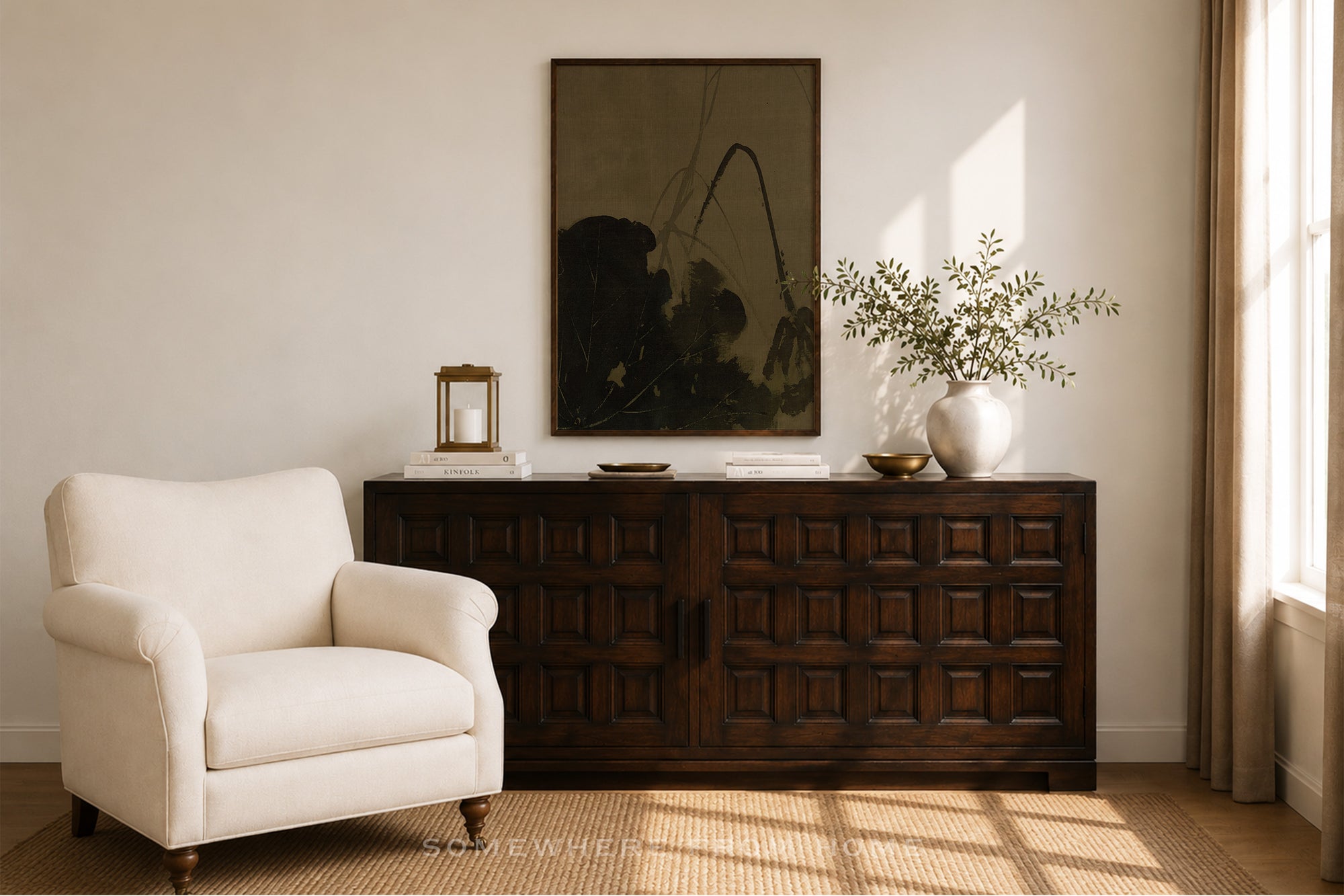 A styled interior scene showing a dark wood sideboard or credenza in a room, with a cream upholstered armchair beside it, a framed dark artwork on the wall, a small lantern, books, a vase with branches, and warm sunlight streaming in from a window.