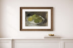 A framed still life art print depicting green and yellow apples on a white plate, mounted on a white wall above a white console table with a small gold bowl.