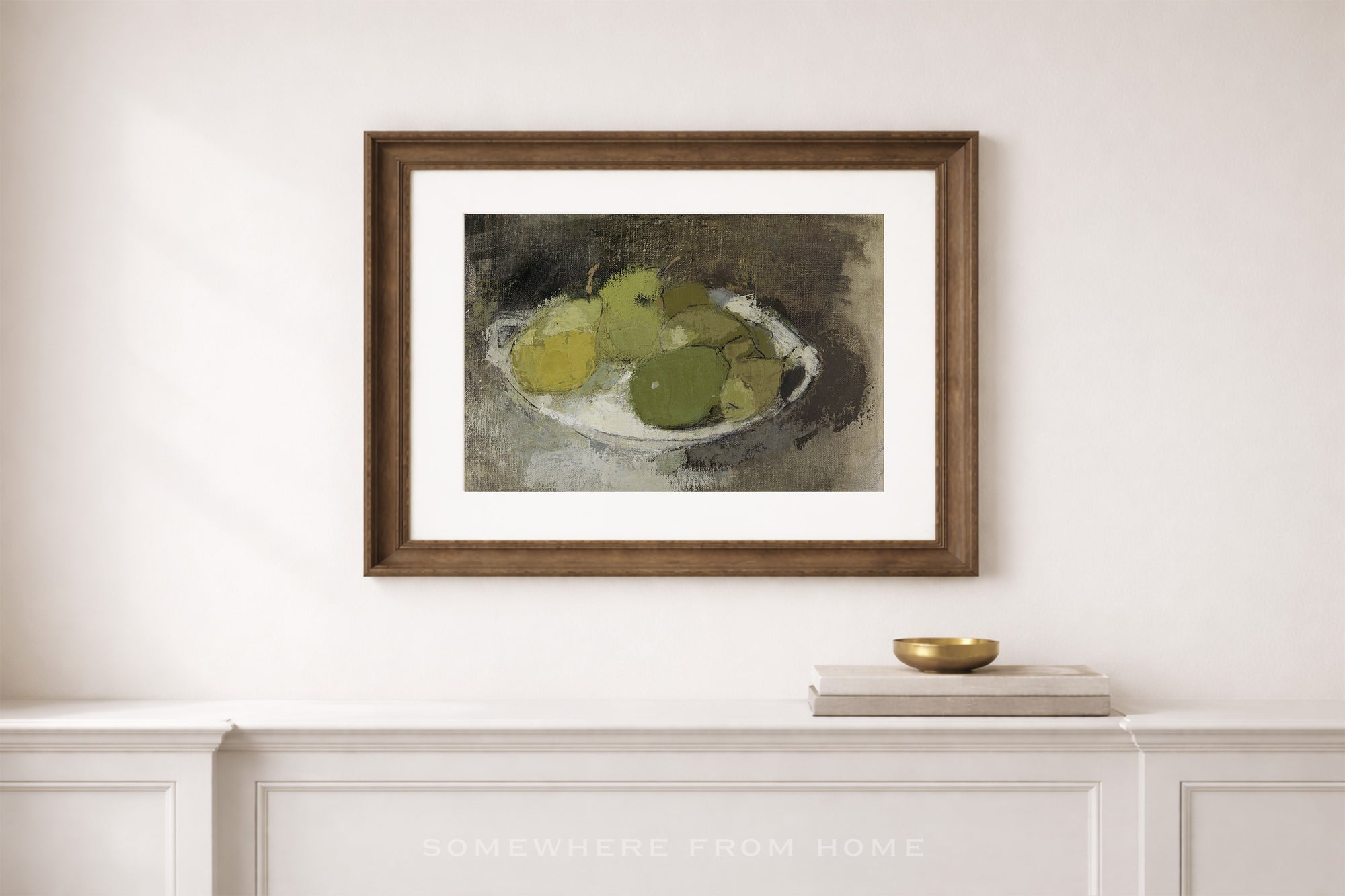 A framed still life art print depicting green and yellow apples on a white plate, mounted on a white wall above a white console table with a small gold bowl.
