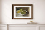A framed still life art print depicting green and yellow apples on a white plate, mounted on a white wall above a white console table with a small gold bowl.